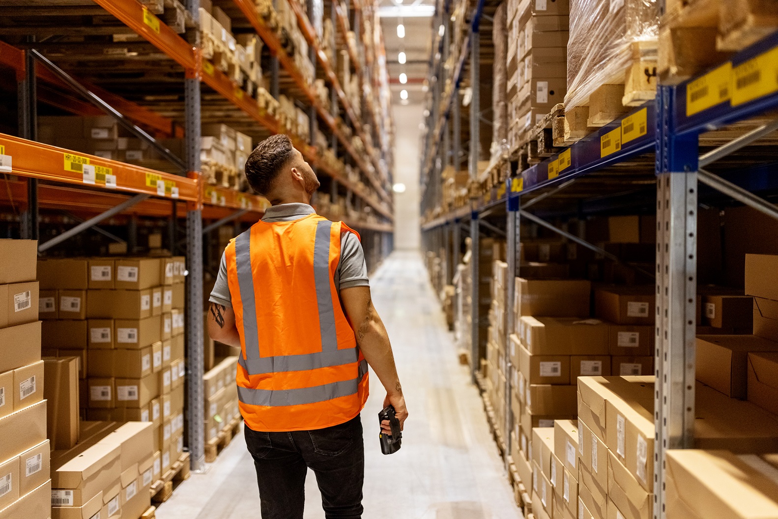 A man confidently strides through a bustling distribution warehouse, surrounded by stacks of pallets and shelves filled with goods. The warehouse is a hive of activity, with workers efficiently moving products and machinery humming in the background. The man's purposeful demeanor reflects the value of investing in warehouses and industrial real estate, highlighting their role as crucial components of supply chains and economic infrastructure. The scene underscores the potential for growth and stability in this sector, as businesses rely on warehouses to store and distribute goods, making them attractive investments in the realm of commercial real estate.